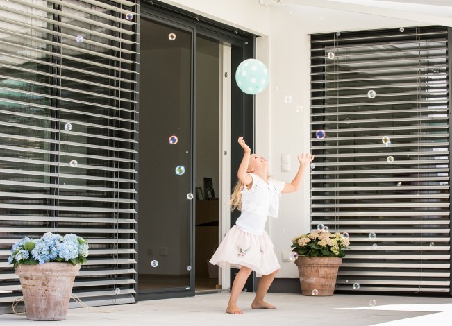 spielendes Mädchen mit einem Luftbaloon vor großen Fenstern mit Raffstores