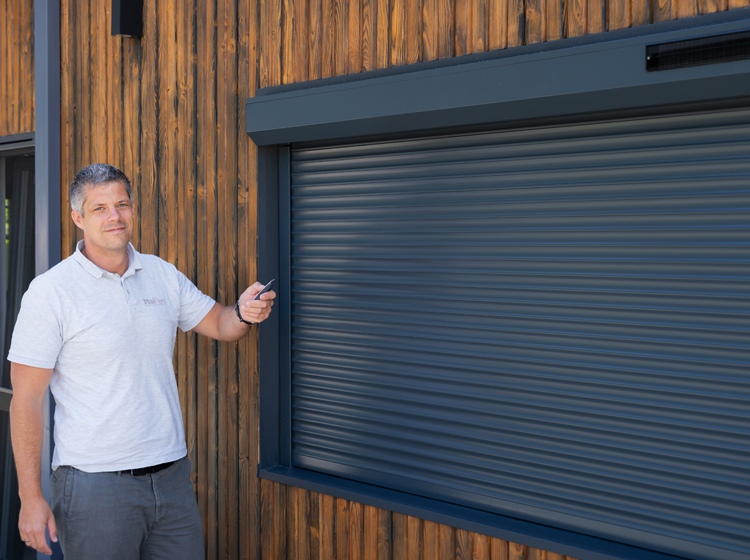 MaiFen-GmbH Fenster- und Sonnenschutztechnik Ing. Maislinger Geschäftsführer von MaiFen GmbH Experte für Sonnenschutz und Insektenschutz vor einem schwarzen eingebauten Solar-Rollladen mit einer Fernbedienung in der Hand